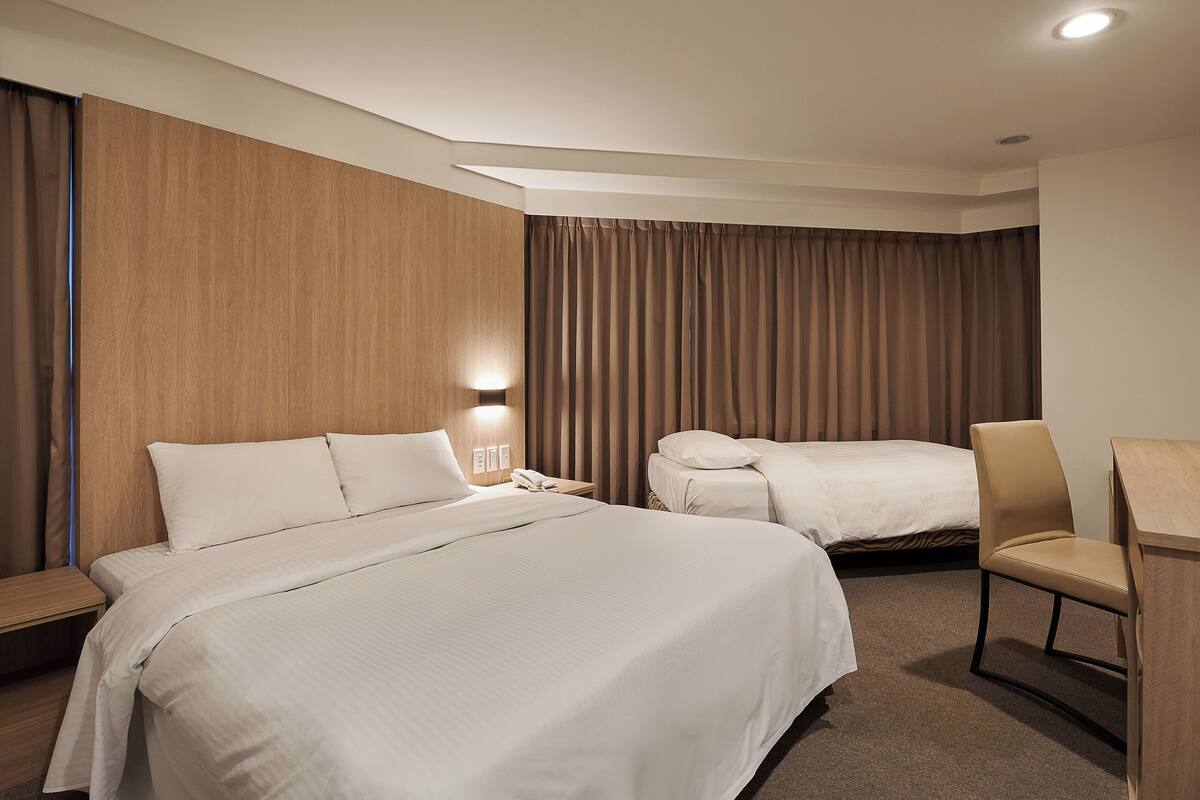 Quarto de hotel com duas camas, uma escrivaninha, uma cadeira e uma mesa de cabeceira com abajur.