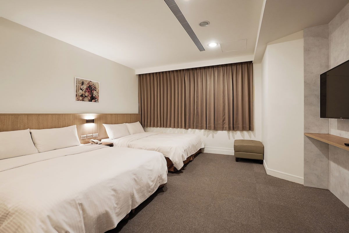 Quarto de hotel com duas camas, uma televisão de tela plana, uma janela grande com cortinas e um pequeno espaço de estar.