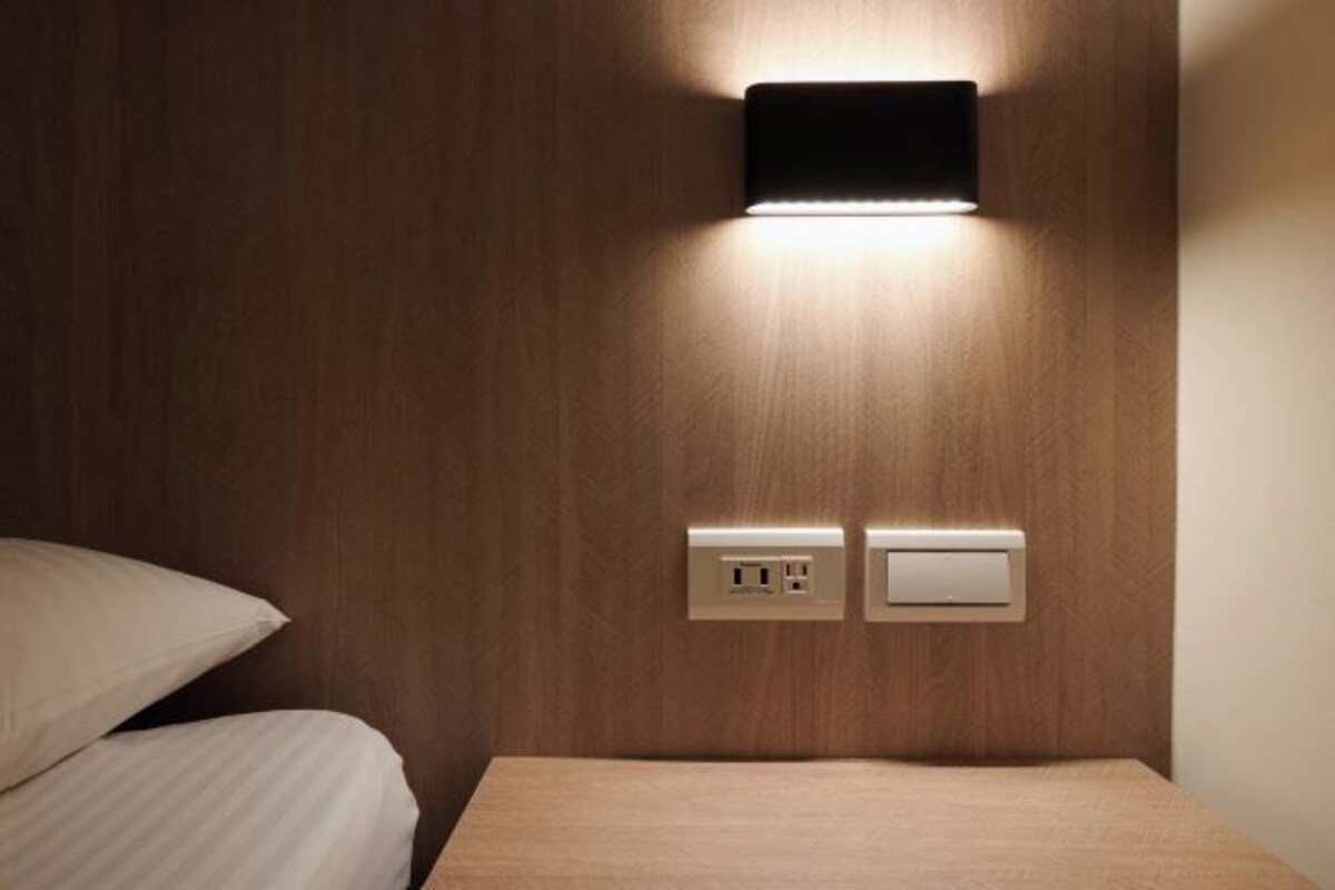 A neatly made bed with a wooden headboard, a wall-mounted light, and two electrical outlets.