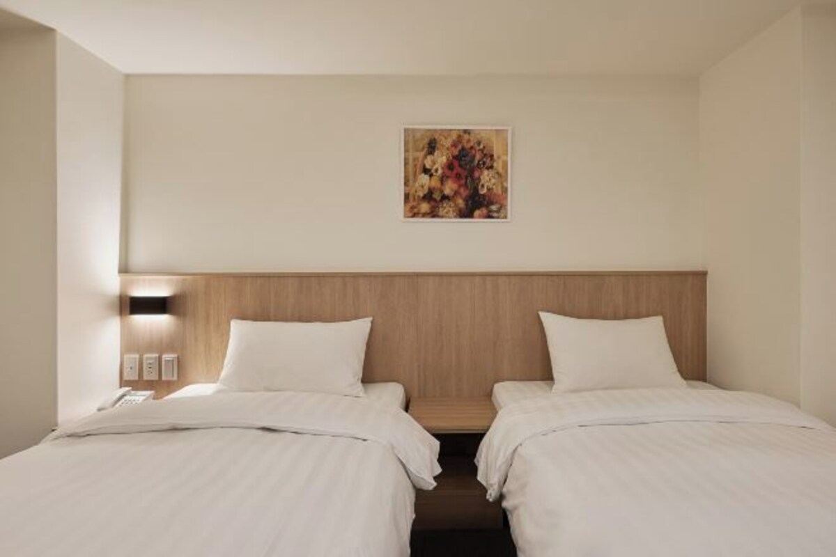 A hotel room with two beds, a wooden headboard, a wall-mounted light, and a painting above the beds.