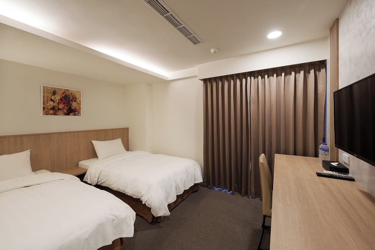 Quarto de hotel com duas camas, uma escrivaninha, televisão e uma janela com cortinas.