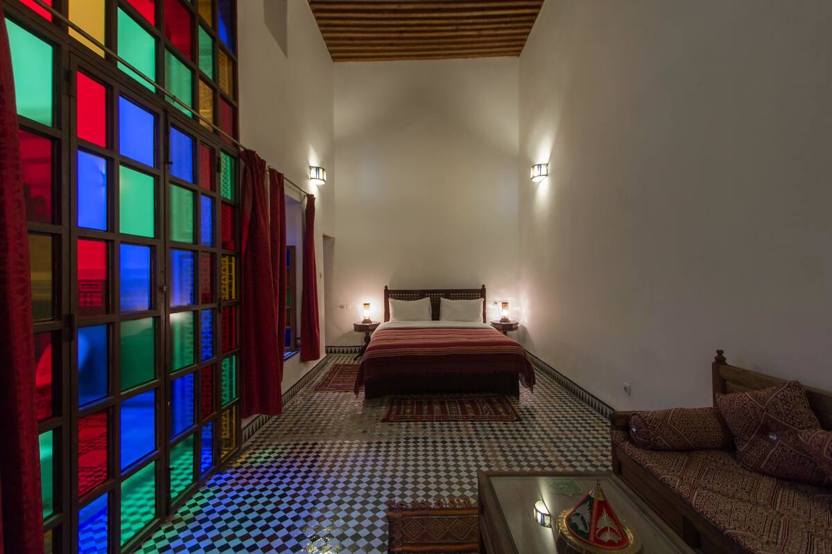 Quarto com piso xadrez, cama com colcha vermelha, sofá de madeira e parede com janelas de vitrais coloridos.
