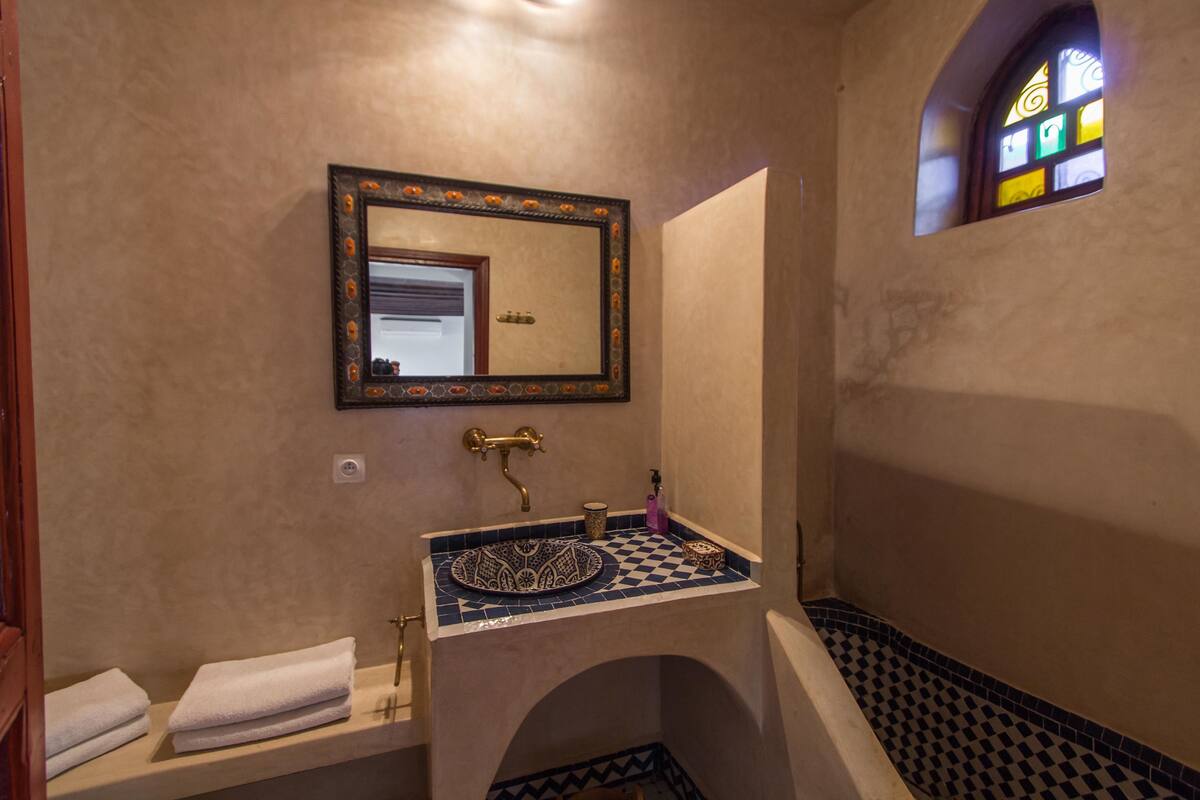 Um banheiro com pia de azulejo quadriculado, um espelho grande e uma pequena janela com vitrais.