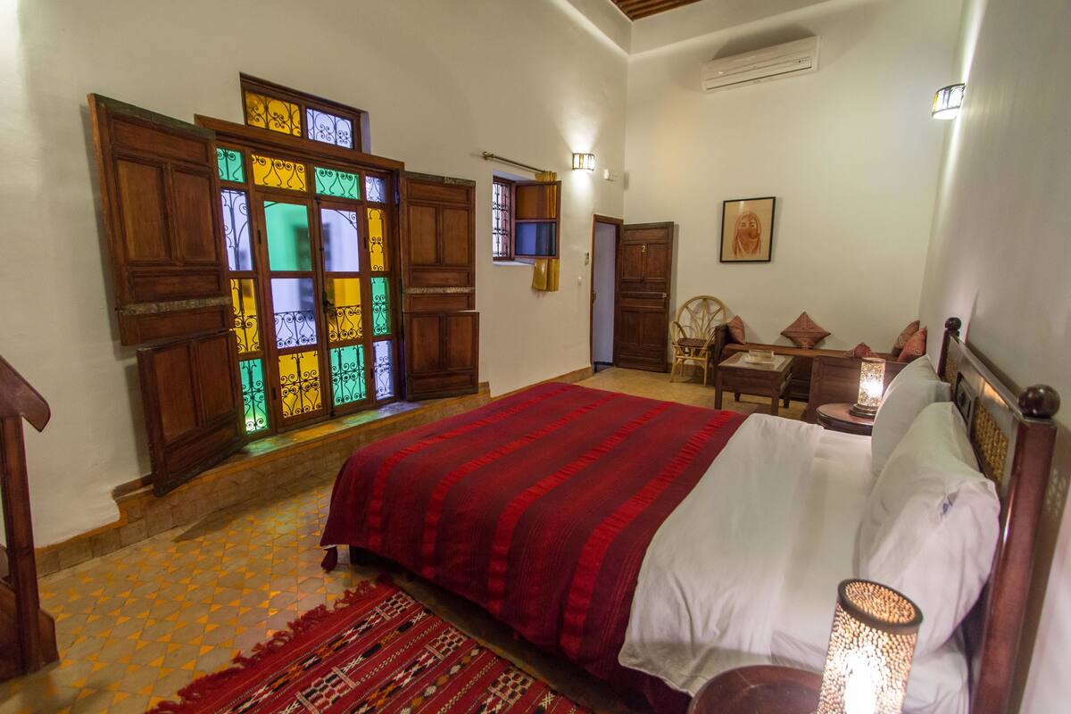 Quarto com uma cama grande, uma porta de madeira com vitrais e uma mesa pequena com abajur.