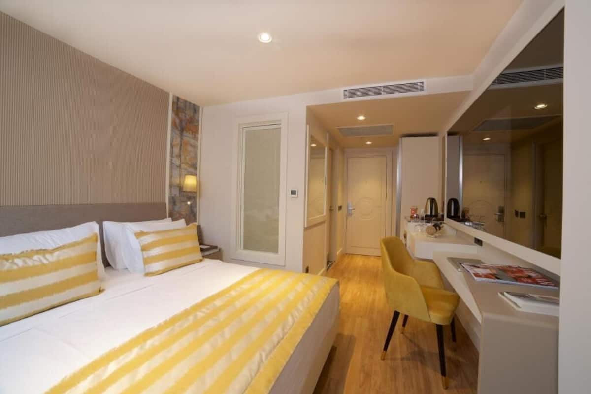 A modern hotel room with a large bed, a sitting area with a chair, a desk, and a large mirror.