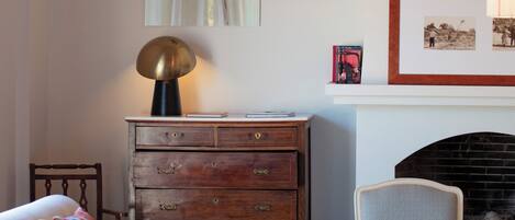 A bedroom with a bed, a wooden dresser, a chair, a table with books, a lamp, and a fireplace.