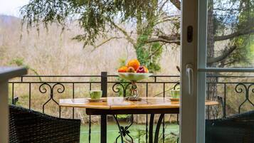 A balcony with a table set for two, a wicker chair, and a view of a body of water and trees.