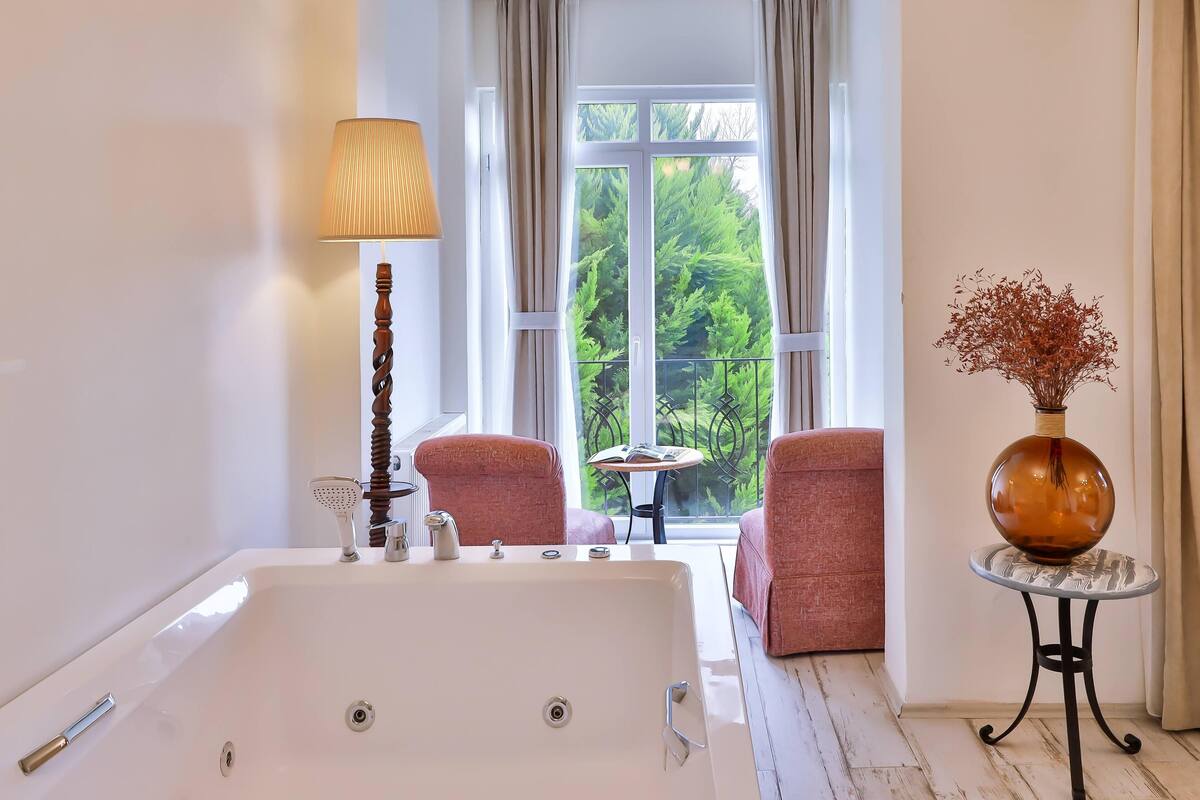 Une salle de bain avec une baignoire, un lampadaire, une chaise, une petite table et un vase rempli de fleurs séchées.
