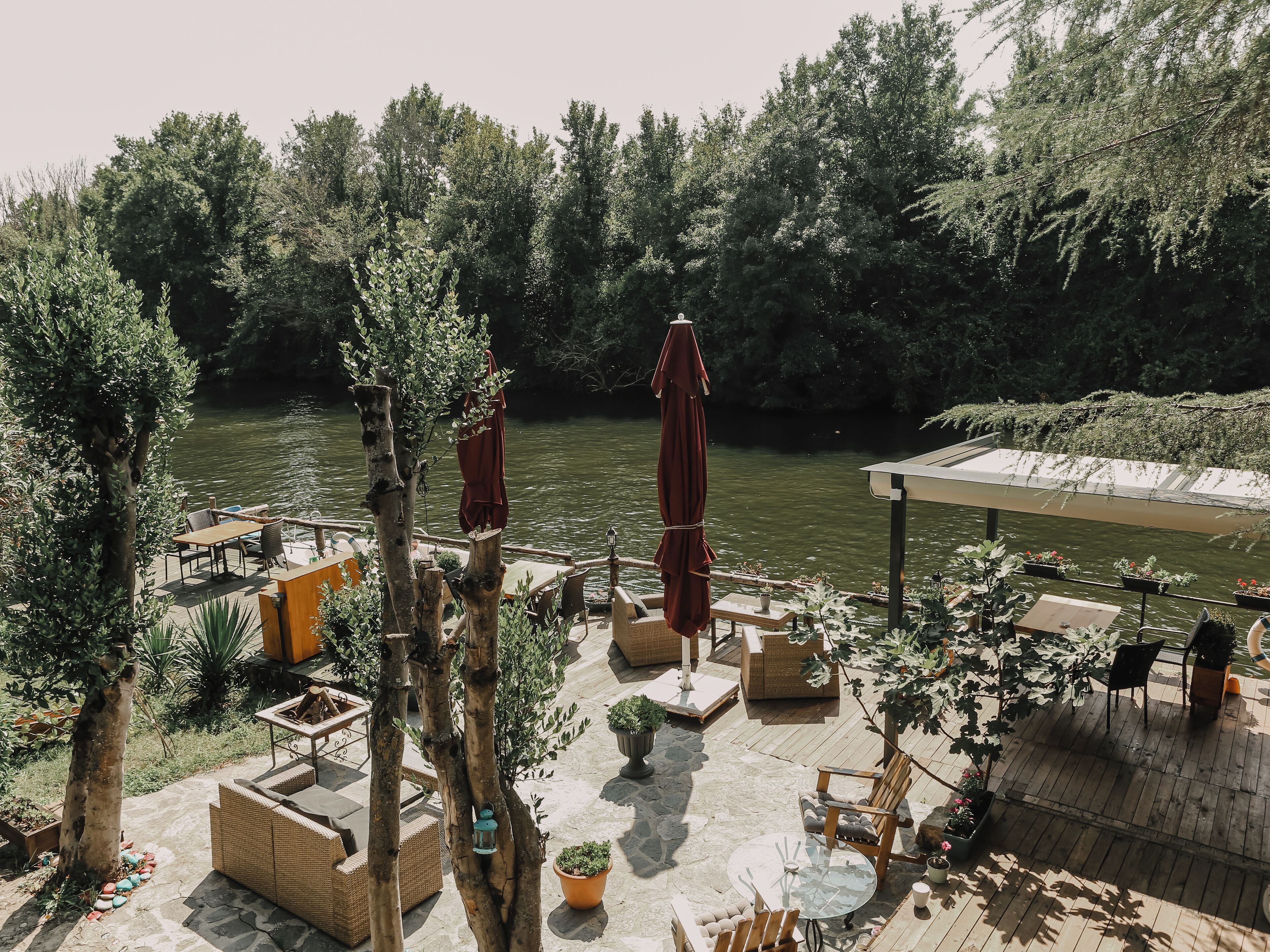 Un patio junto al río con mesas al aire libre, un espacio cubierto y abundante vegetación.