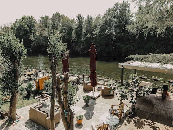 Un patio junto al río con mesas al aire libre, un espacio cubierto y abundante vegetación.