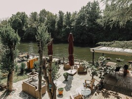 A riverside patio with outdoor seating, a covered area, and lush greenery.