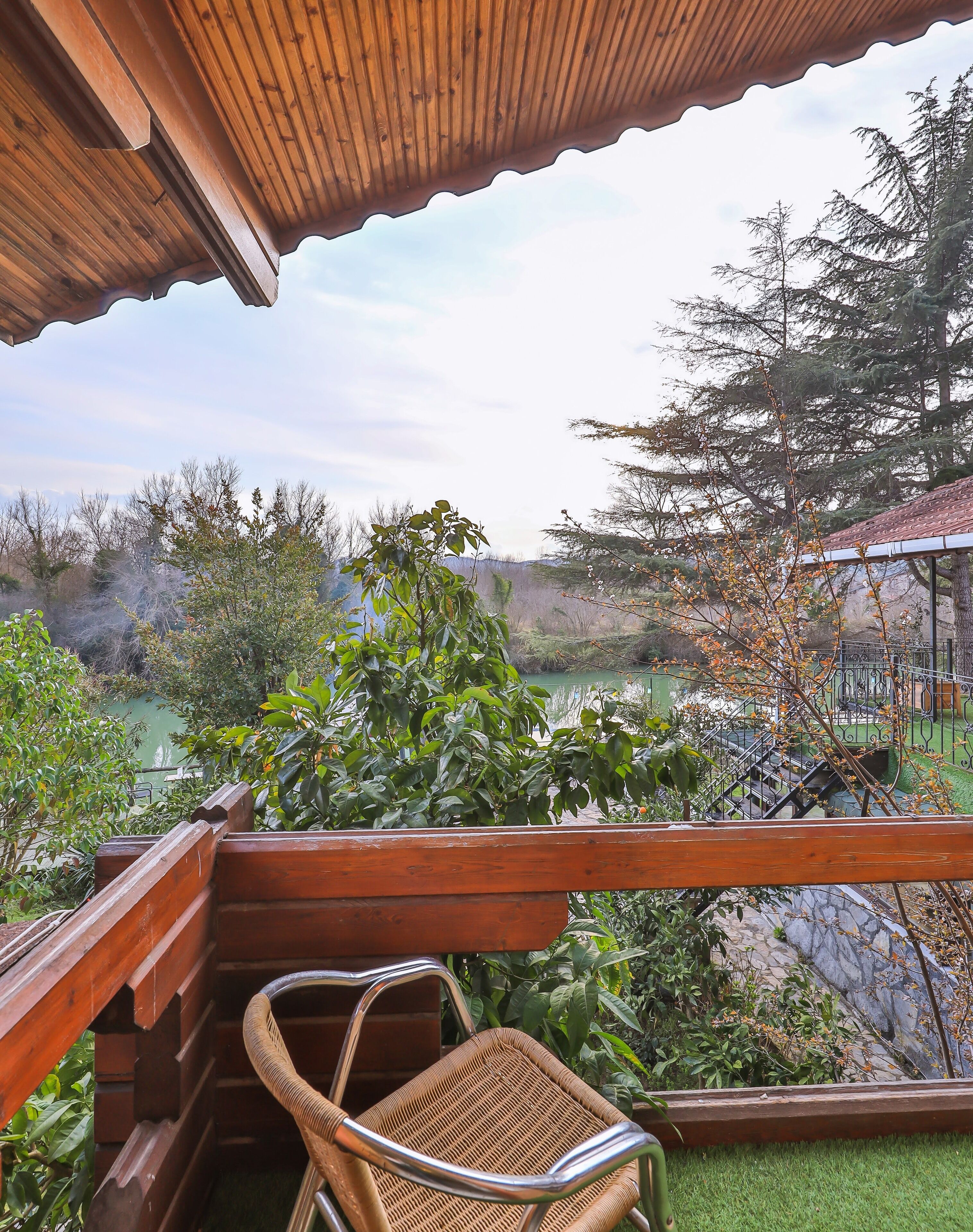 Un balcón de madera con una silla de mimbre, con vista a un jardín con un estanque y árboles.