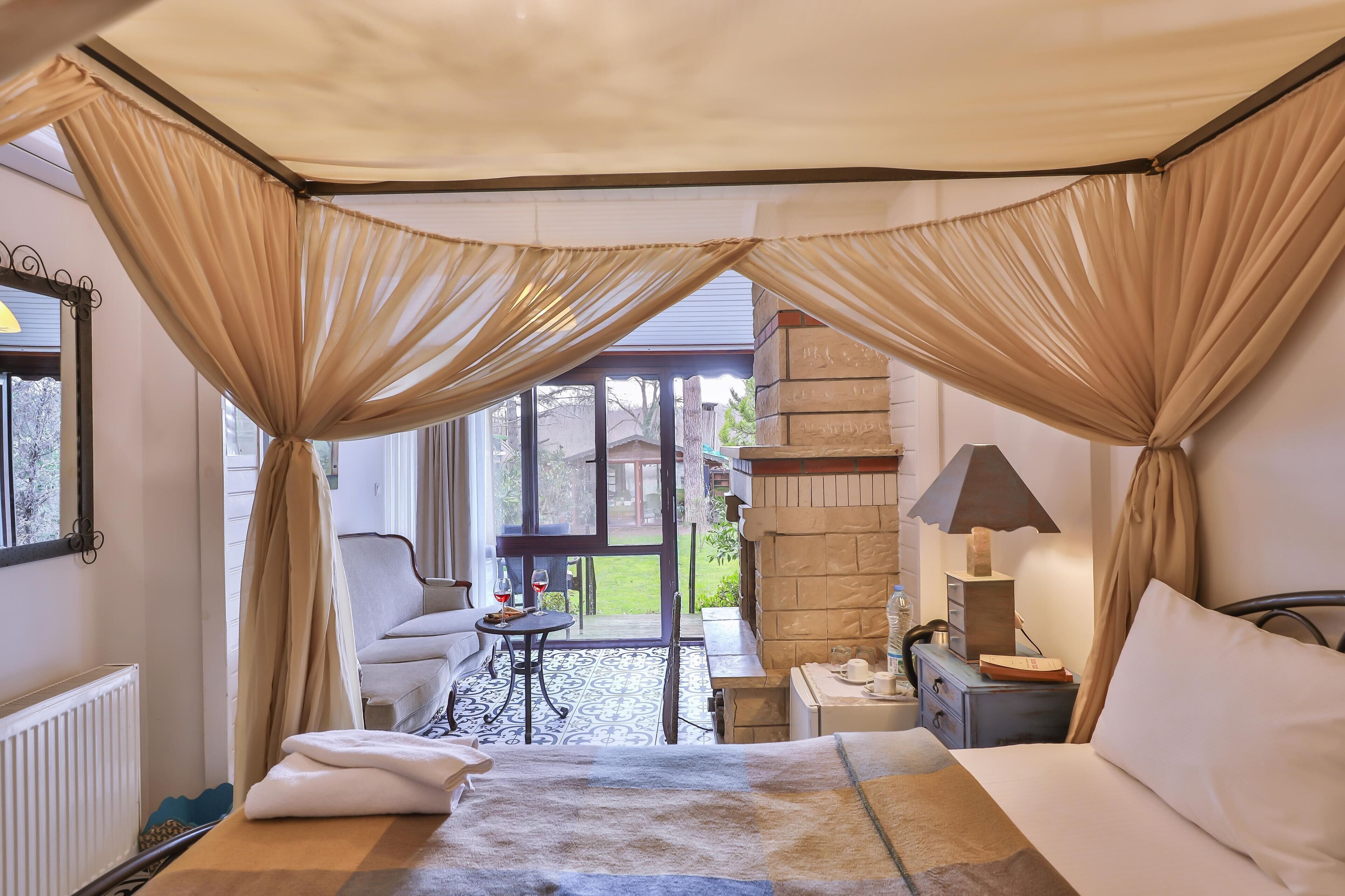 Quarto com cama de dossel, área de estar com mesa e cadeiras, pequena escrivaninha com luminária, e vista para o exterior.