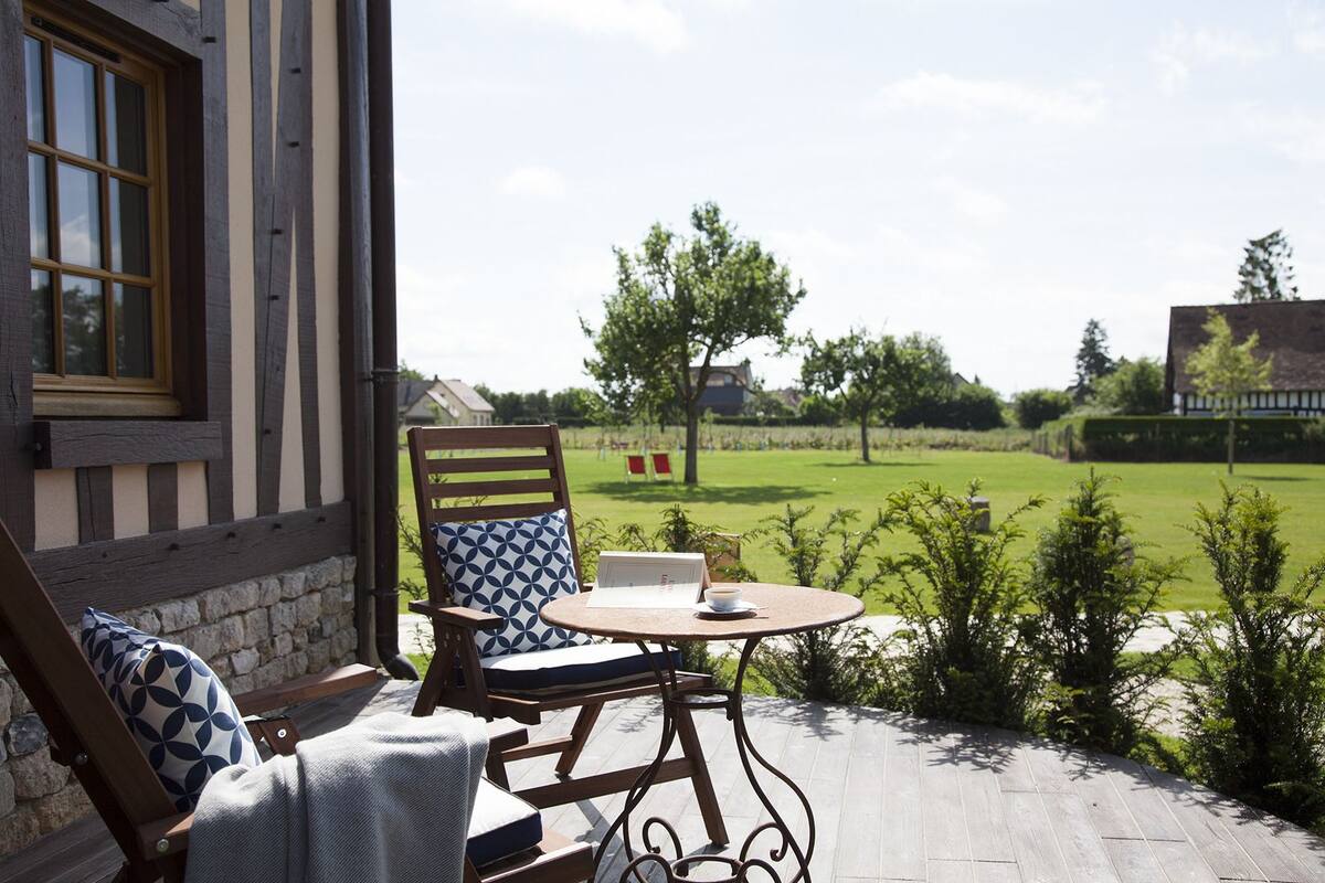 Une terrasse en bois avec une table et des chaises, donnant sur une zone herbeuse avec des arbres et un bâtiment en arrière-plan.