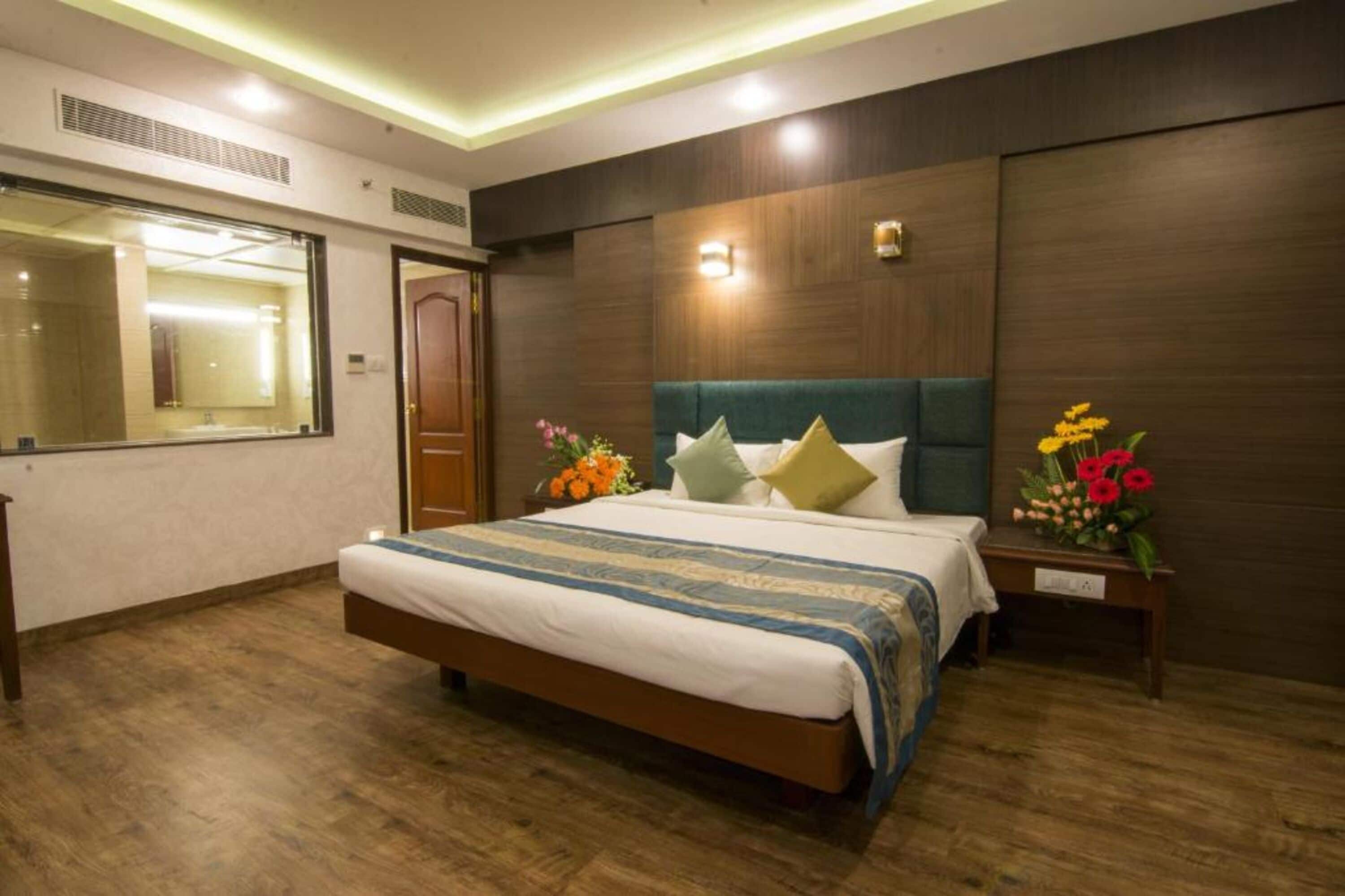 A hotel room with a large bed, wooden flooring, a mirror, and a nightstand with flowers.
