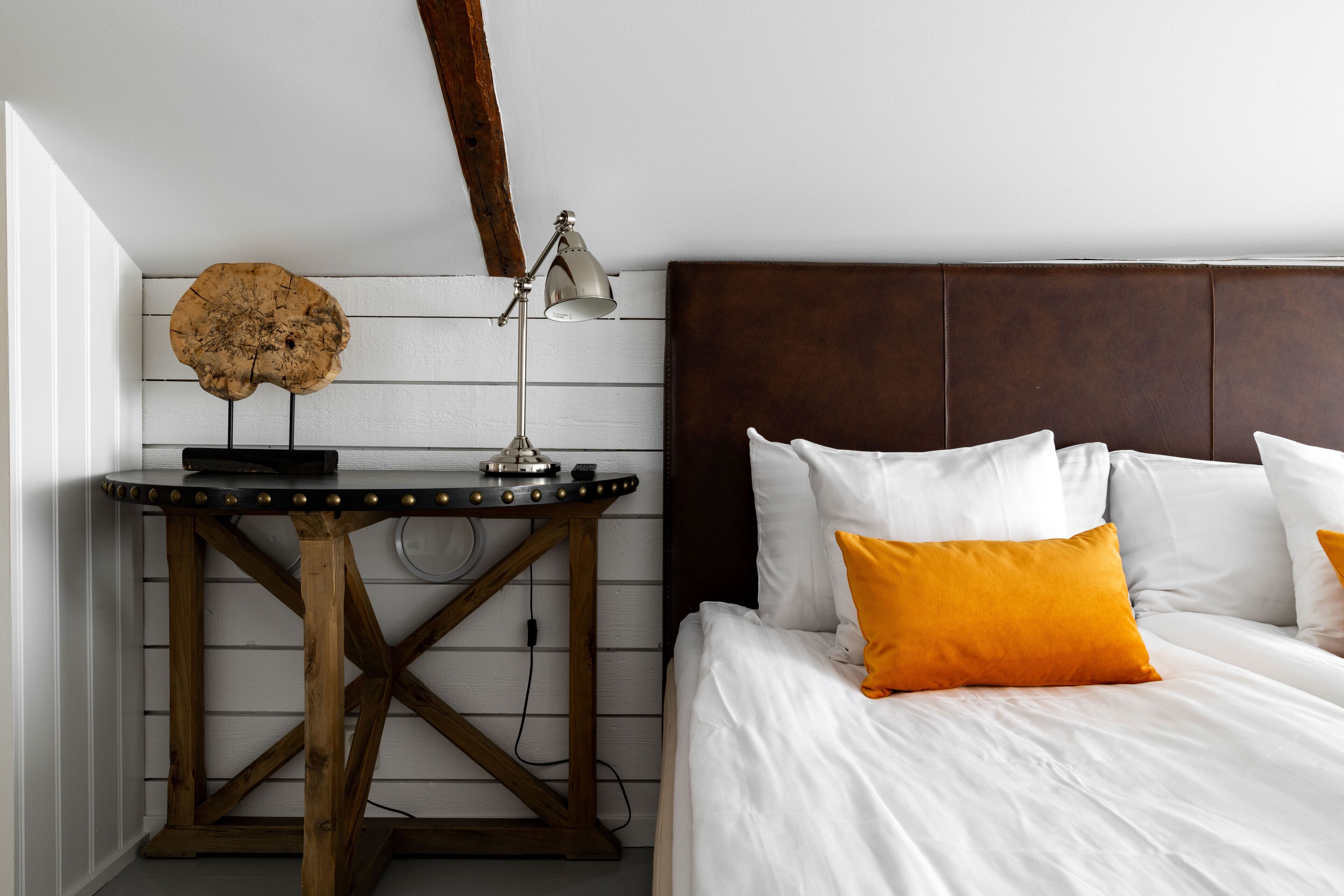 Un dormitorio con una mesita de noche de madera, una lámpara, una cama con ropa de cama blanca y un cojín naranja, y un cabecero de madera.