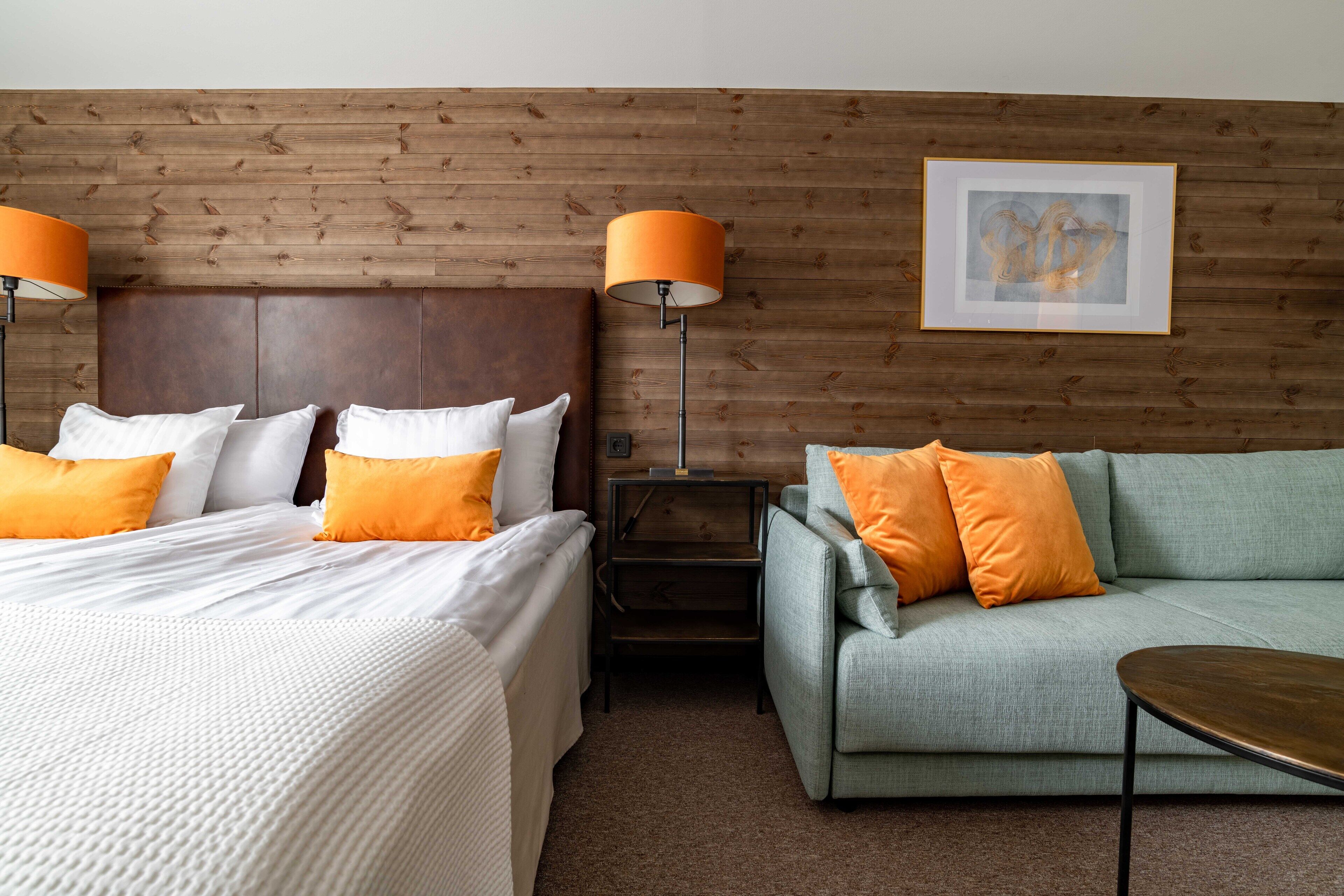 Ett hotellrum med en sänggavel i trä, två sängar med vita och orange sängkläder, en grön soffa, ett litet bord och en golvlampa.