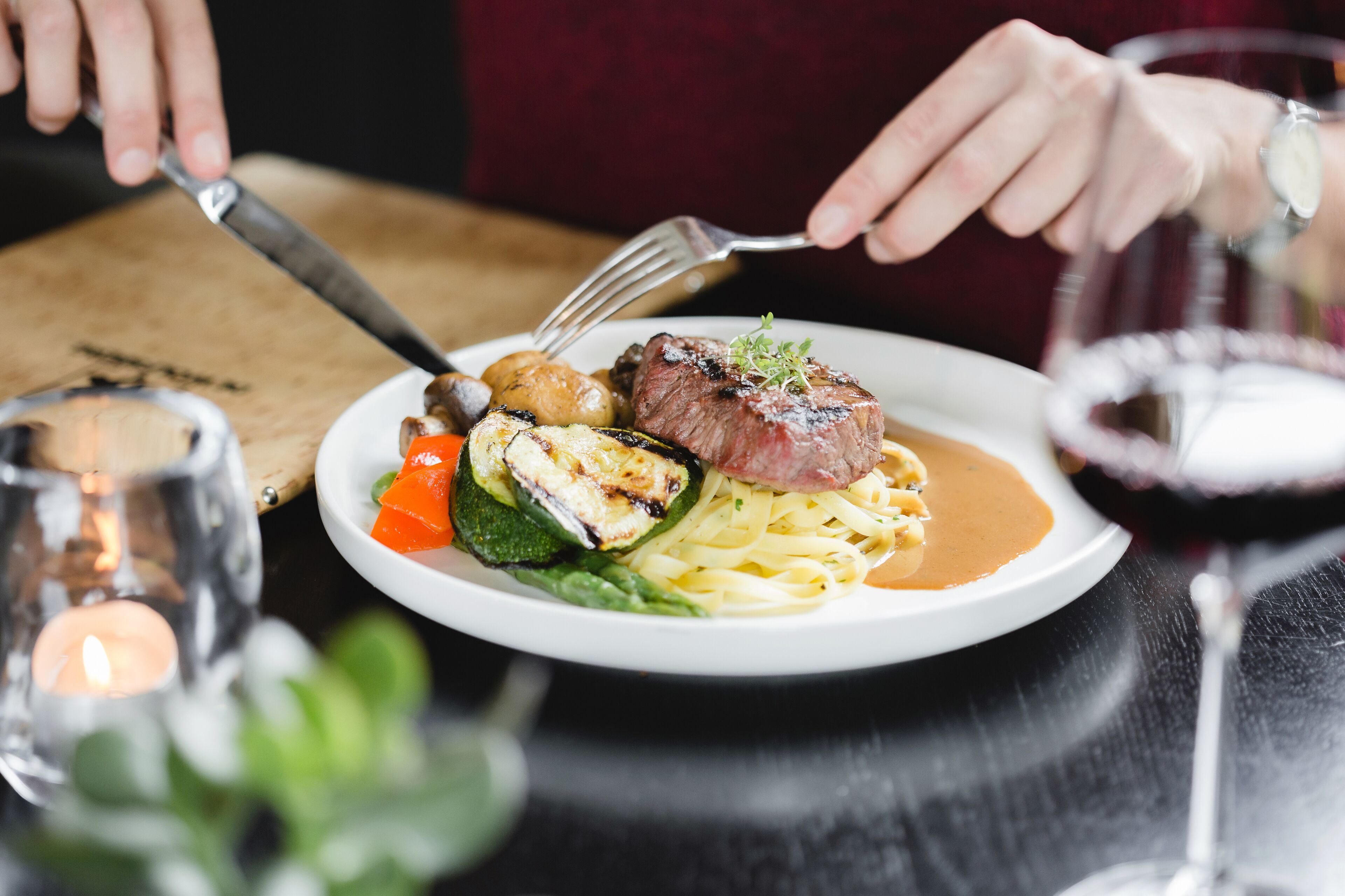 Prato de comida com bife, legumes e massa, acompanhado por um copo de vinho tinto.