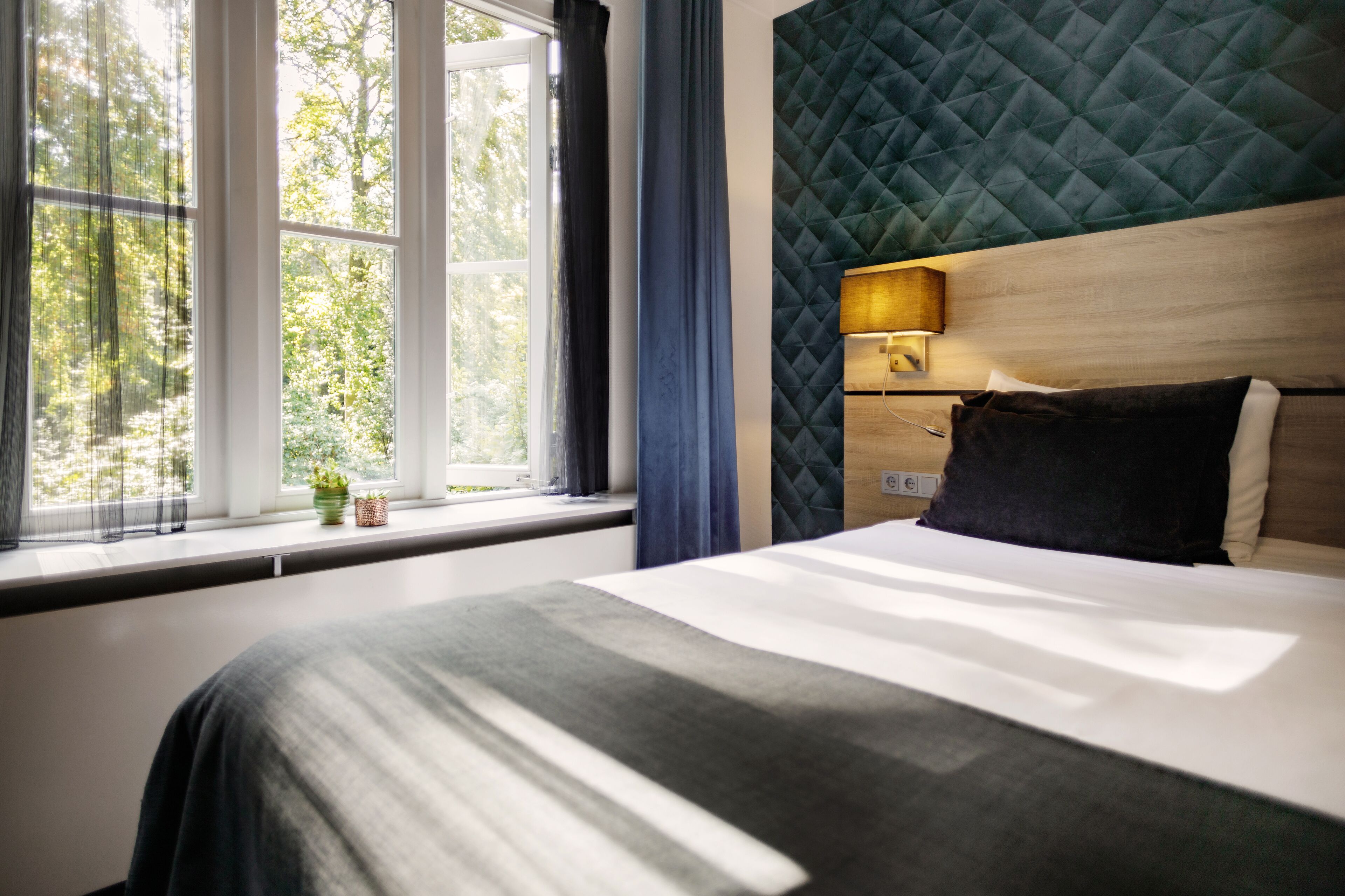Una habitación de hotel moderna con una cama grande, una ventana con cortinas, vistas a zonas verdes y un cabecero acolchado.