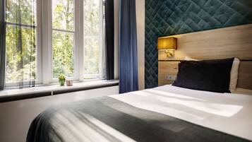 Una habitación de hotel moderna con una cama grande, una ventana con cortinas, vistas a zonas verdes y un cabecero acolchado.