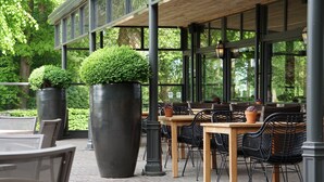 Een terras met tafels en stoelen, grote plantenbakken en een overdekt zitgedeelte met grote ramen.