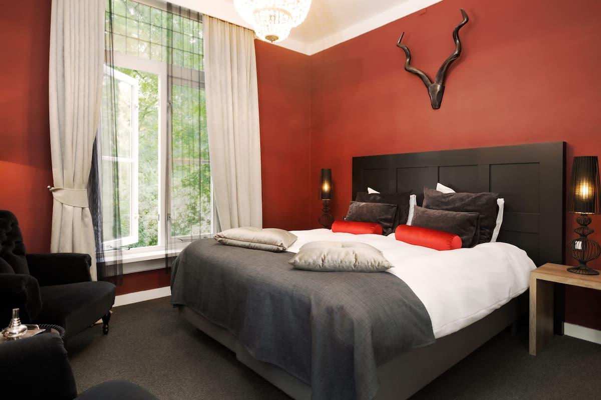 Une chambre à coucher avec un grand lit, une chaise, une table de chevet et une fenêtre avec des rideaux.