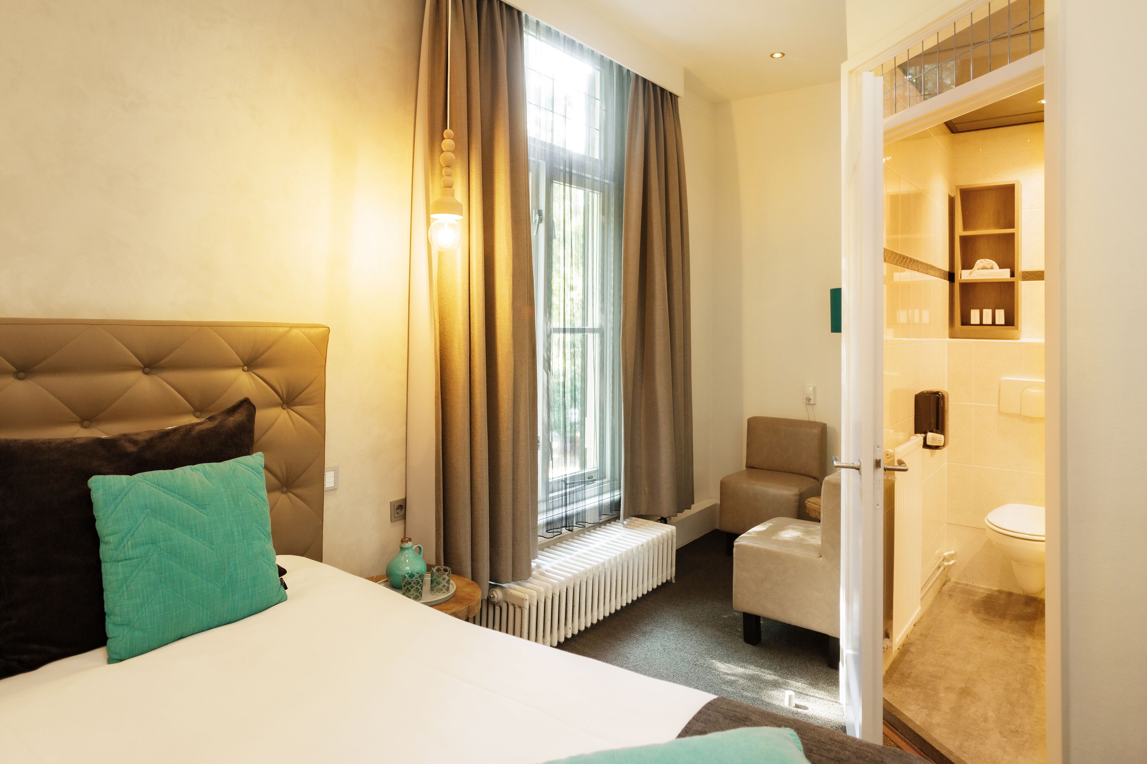 Habitación de hotel con una cama grande, un radiador, una ventana con cortinas, una silla y un baño visible a través de una puerta abierta.