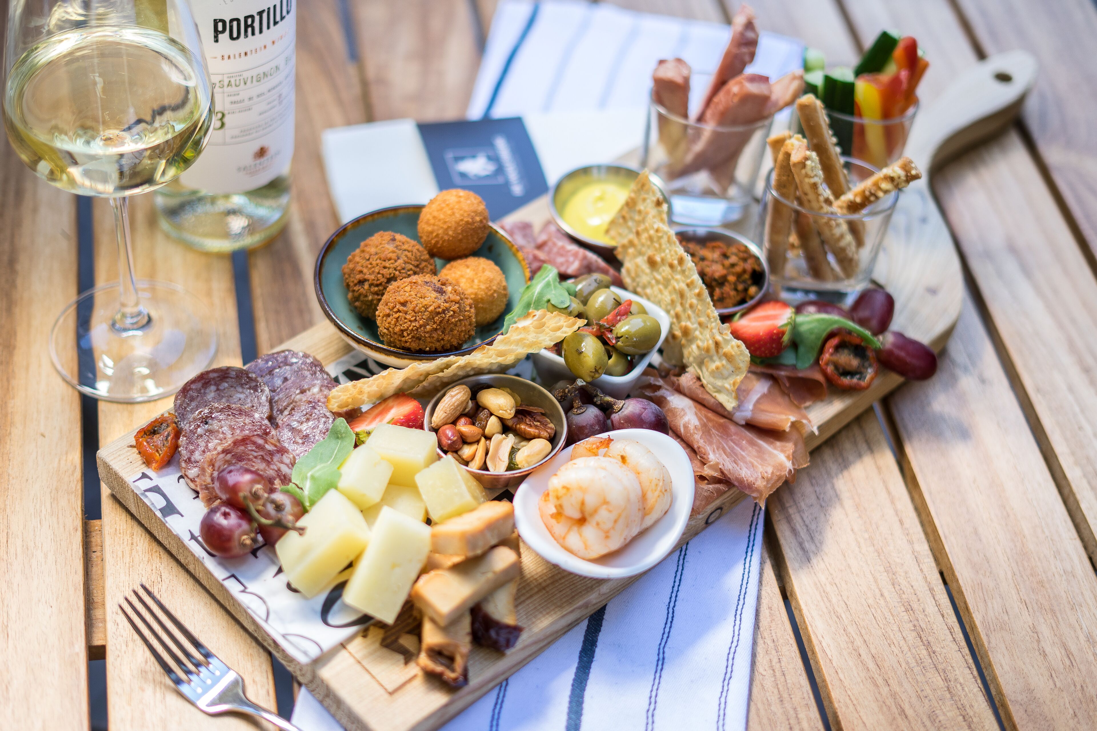 Una tabla de madera con diversos quesos, embutidos, aceitunas y galletas. Se ve también una botella de vino blanco y una copa.