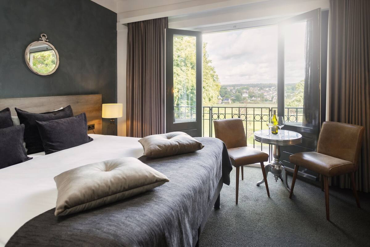Une chambre d’hôtel avec un grand lit, deux chaises, une petite table avec une bouteille et un verre, et un balcon offrant une vue sur la ville.