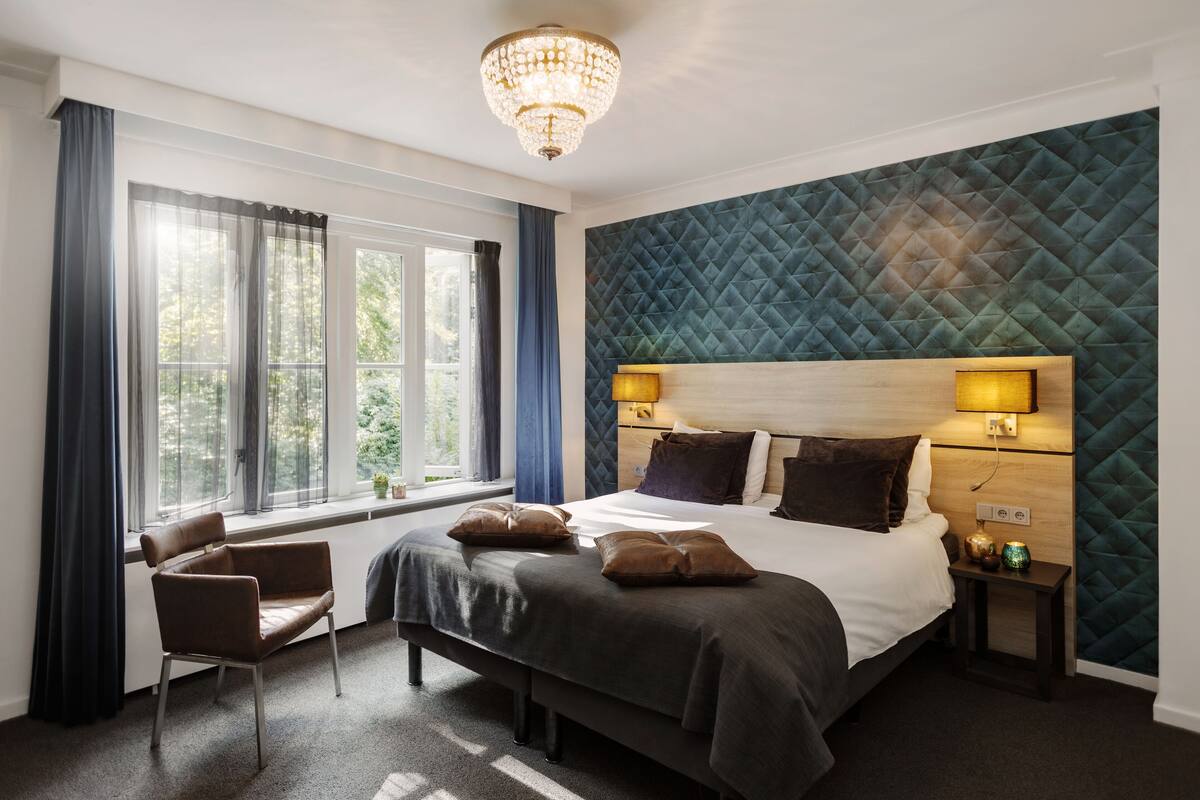 Une chambre d’hôtel moderne dotée d’un grand lit, d’un coin salon avec un fauteuil, d’une fenêtre avec des rideaux et d’un panneau décoratif mural.