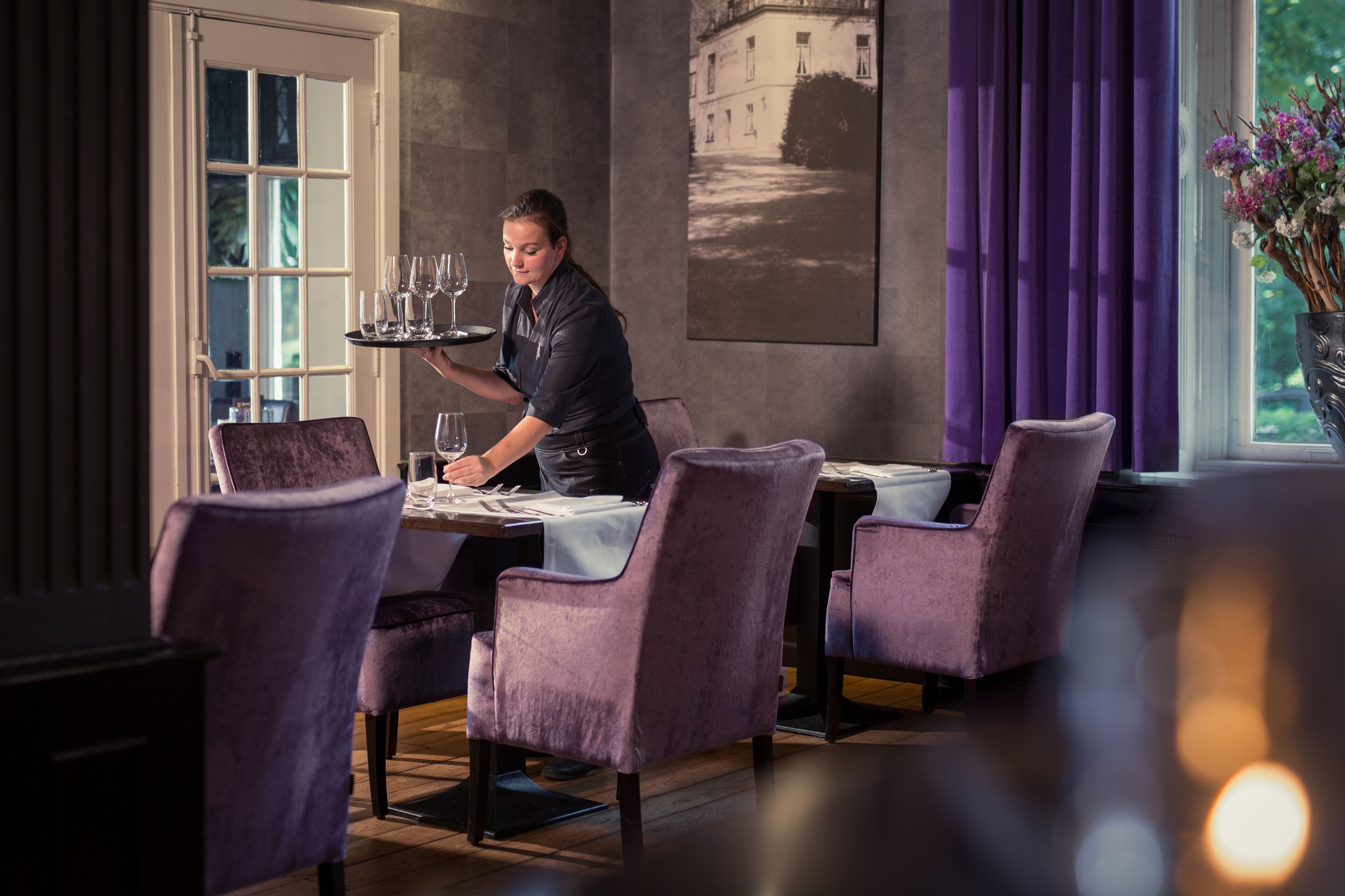 Une salle à manger avec des chaises violettes, un serveur portant un plateau et une table dressée pour deux personnes.