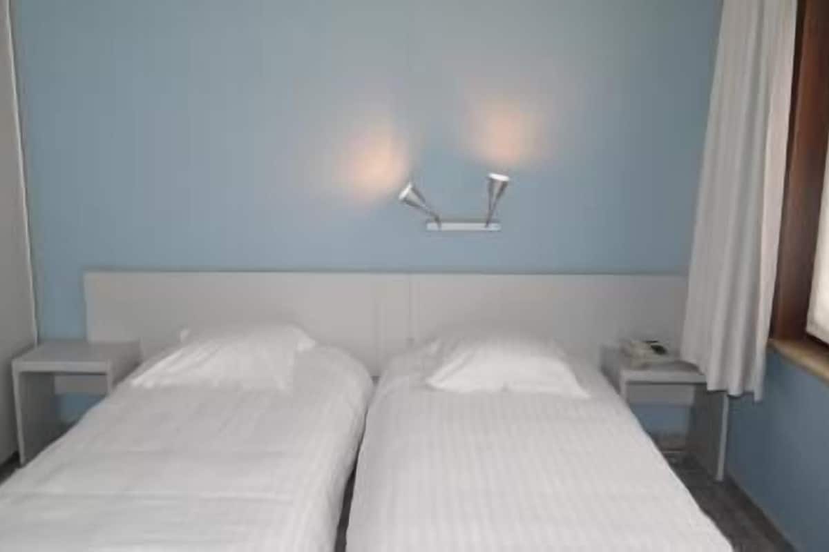 Een tweepersoonsbed met witte beddengoed, twee nachtkastjes en een wandlamp.