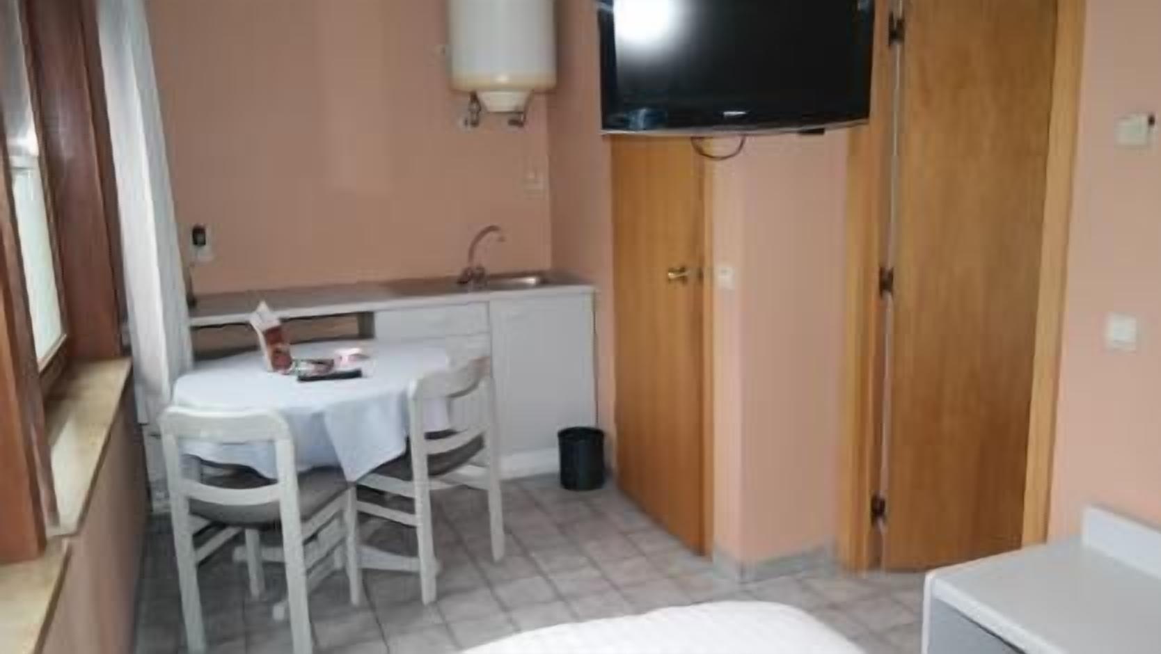 Una habitación pequeña con una cama individual, una mesa con sillas, un televisor en la pared y una puerta de madera.