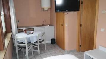Una habitación pequeña con una cama individual, una mesa con sillas, un televisor en la pared y una puerta de madera.