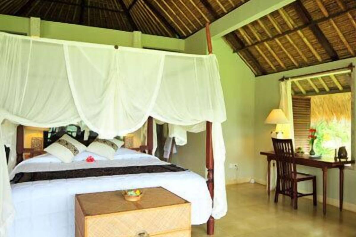 A room with a bed under a canopy, a wooden nightstand, a desk with a chair, and a window with blinds.