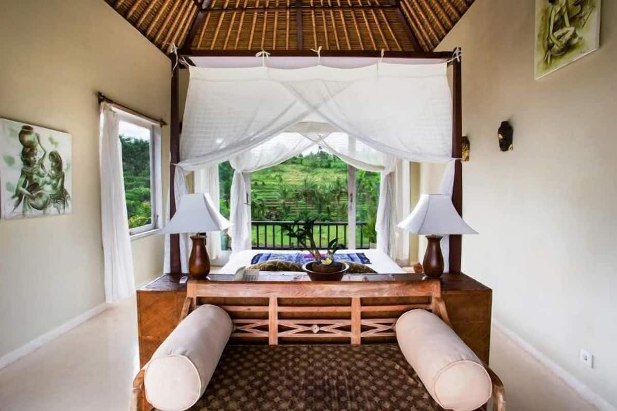 A bedroom with a canopy bed, two bedside lamps, a view of a lush garden, and a wooden headboard.