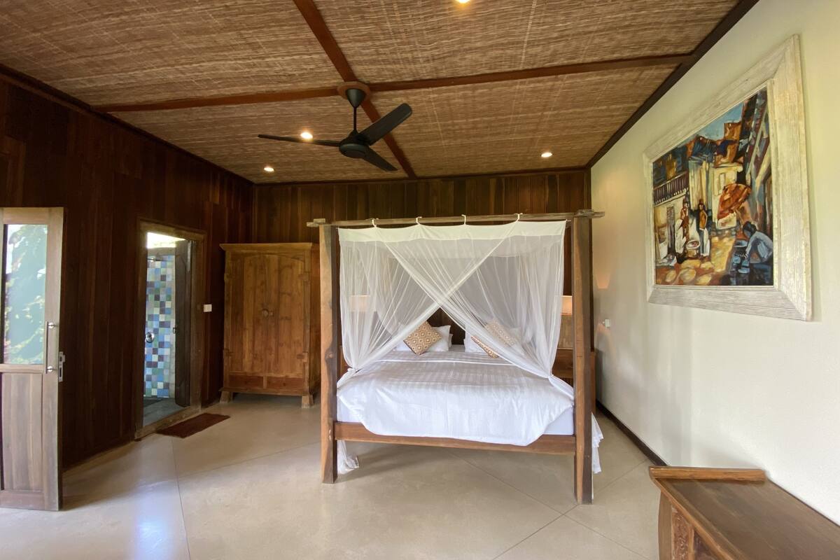 Una cama con dosel de madera y mosquitera, un ventilador de techo y un cuadro en la pared.