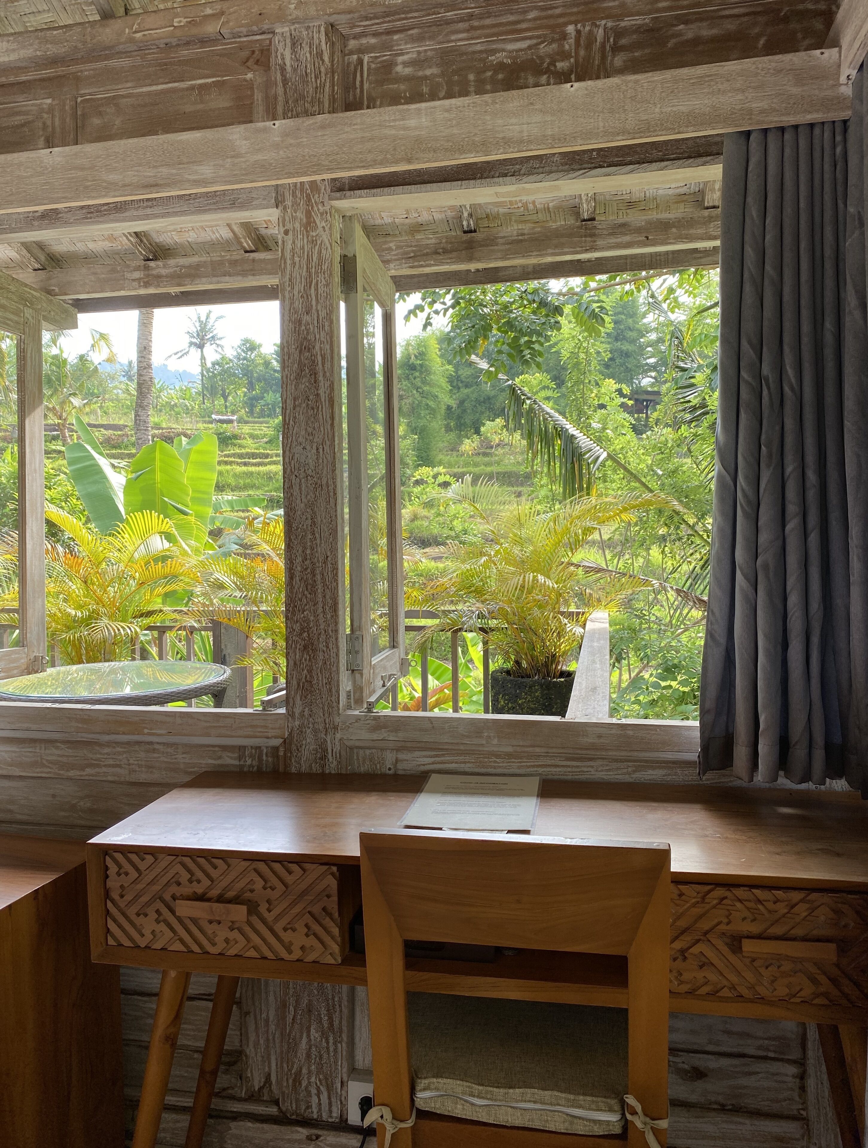 A wooden desk with a chair, a window with a view of a lush garden, and a curtain on the right.