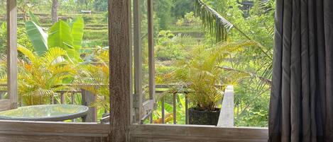 A wooden desk with a chair, a window with a view of a lush garden, and a curtain on the right.