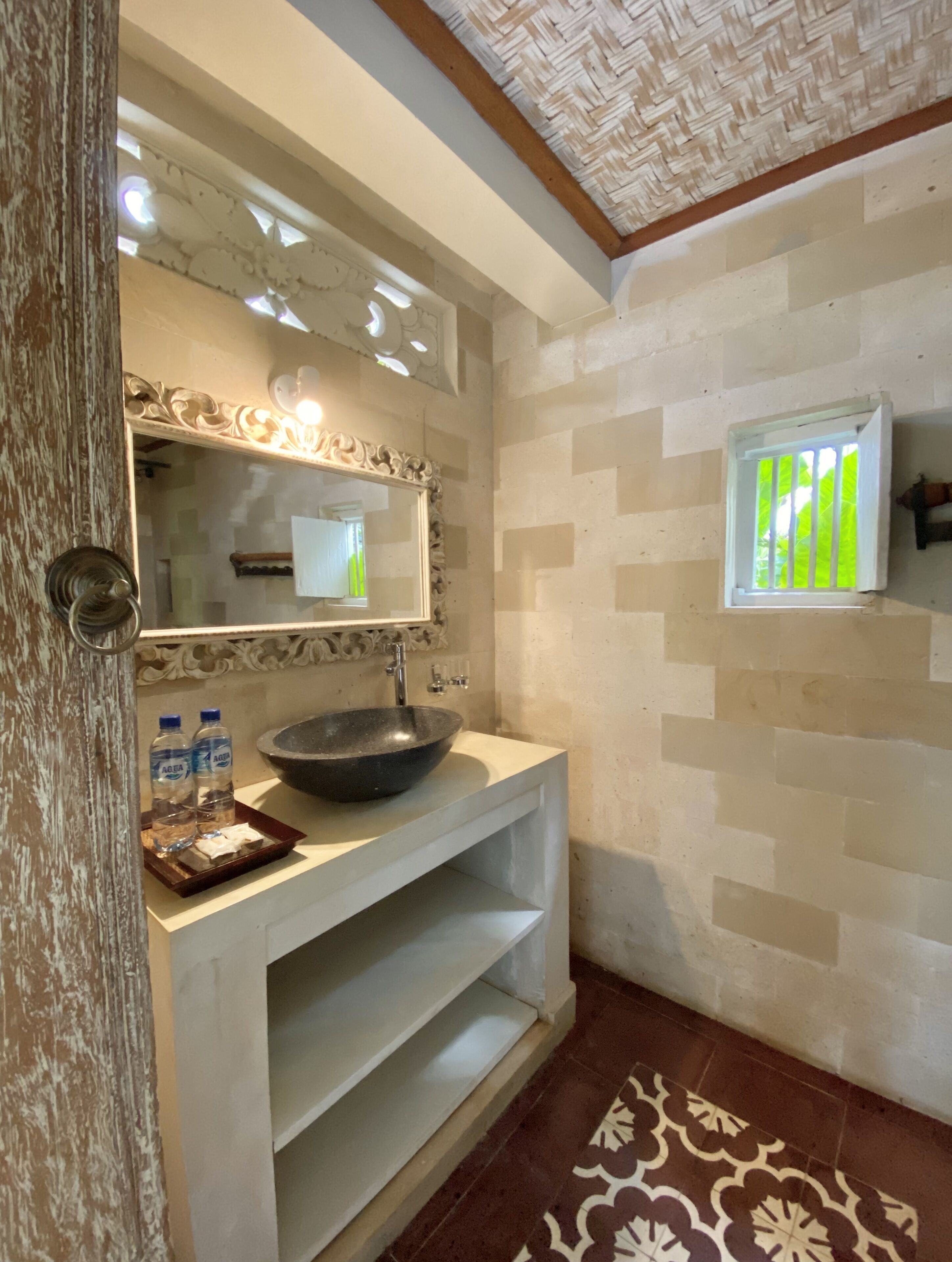 A bathroom with a large mirror, a vessel sink, two bottles of water, a small window, and patterned floor tiles.