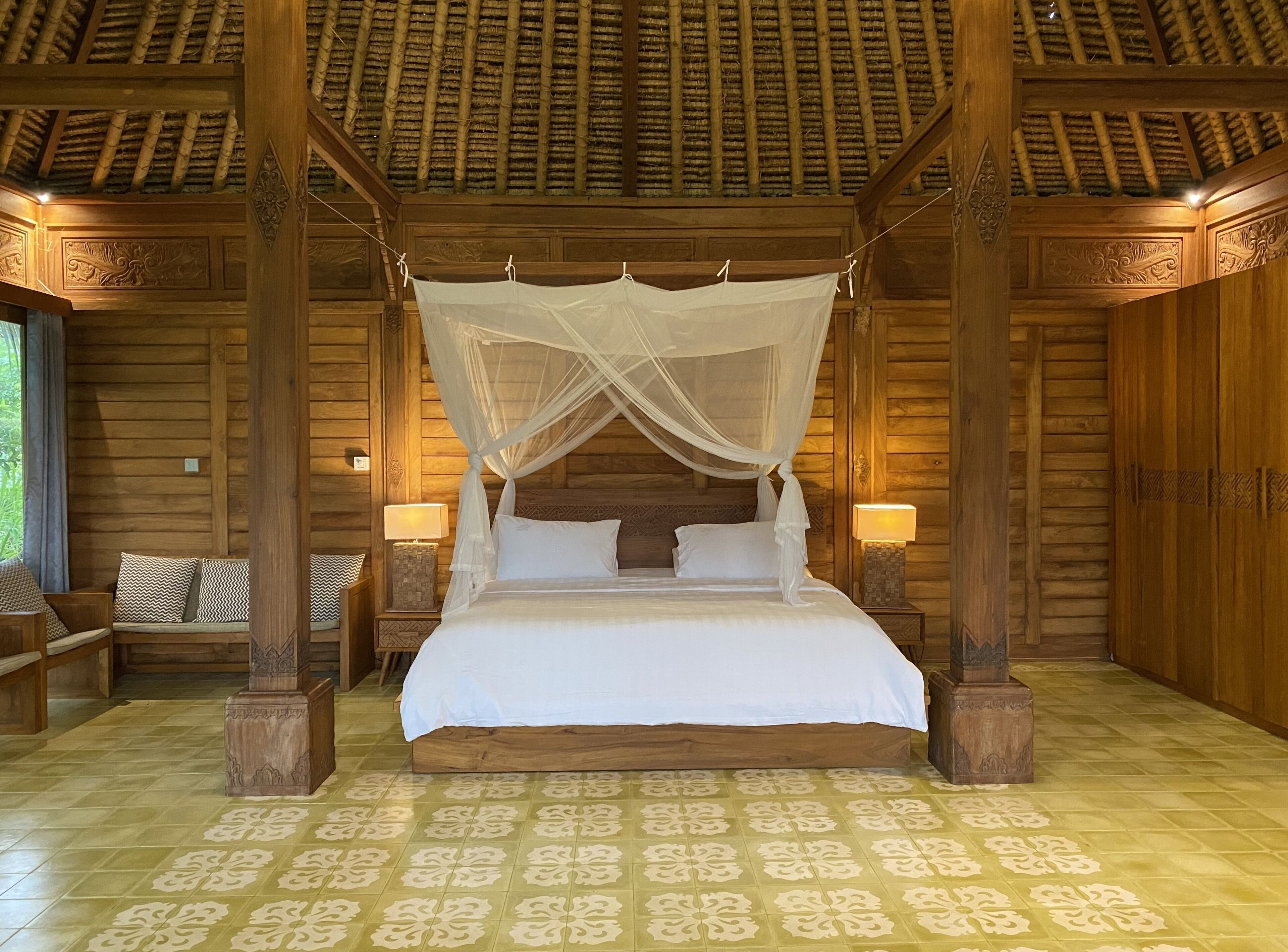 A wooden four-poster bed with a canopy, flanked by nightstands with lamps, in a room with patterned floor tiles and wooden walls.