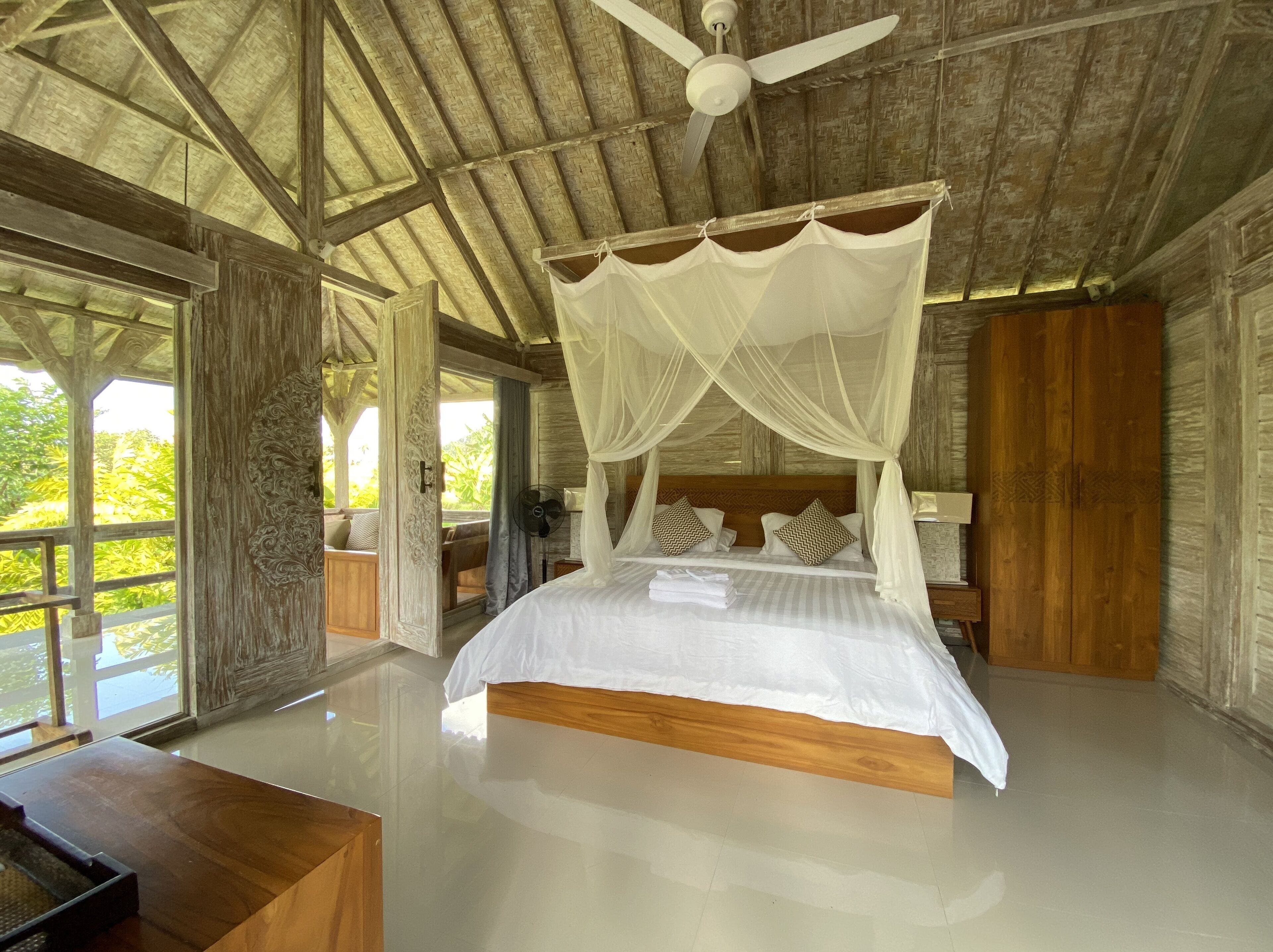 A spacious bedroom with a four-poster bed, a ceiling fan, and a view of lush greenery outside.