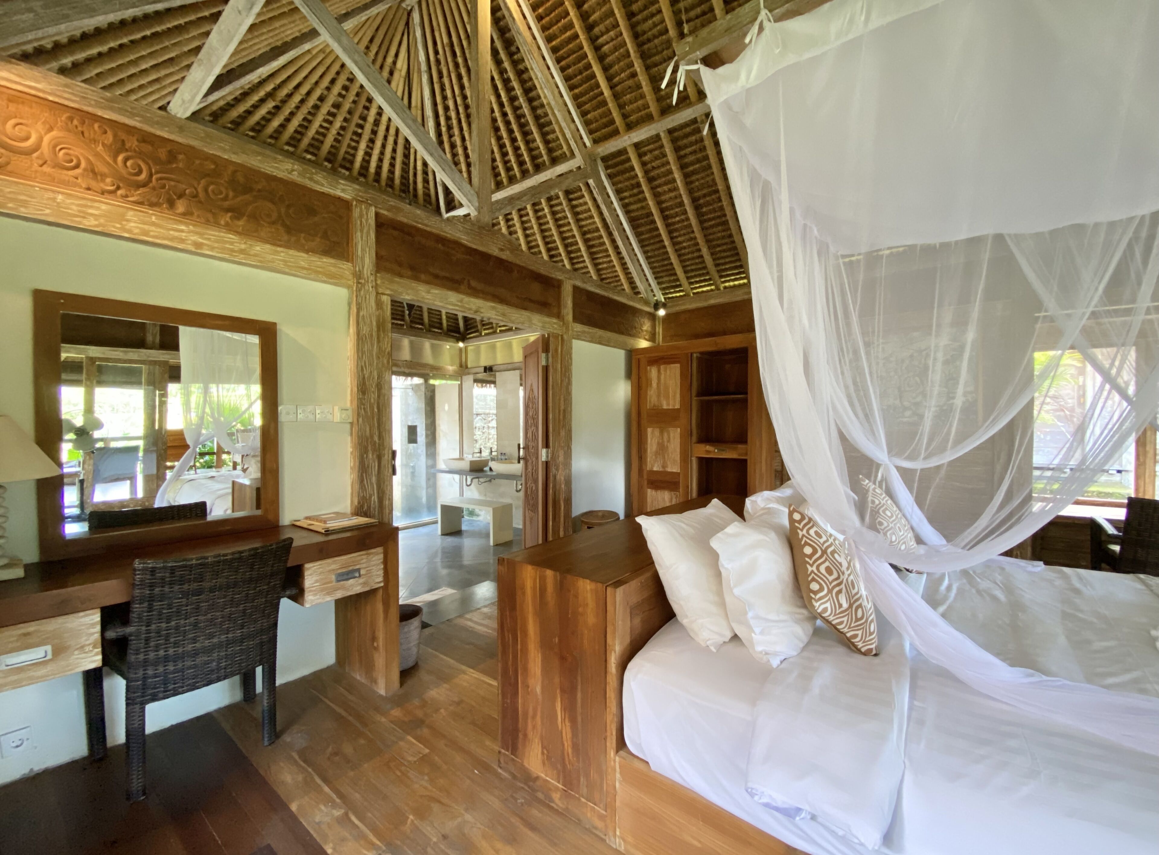 A bedroom with a wooden ceiling, a bed with white bedding, a bedside table, a chair, a desk, and a large mirror.