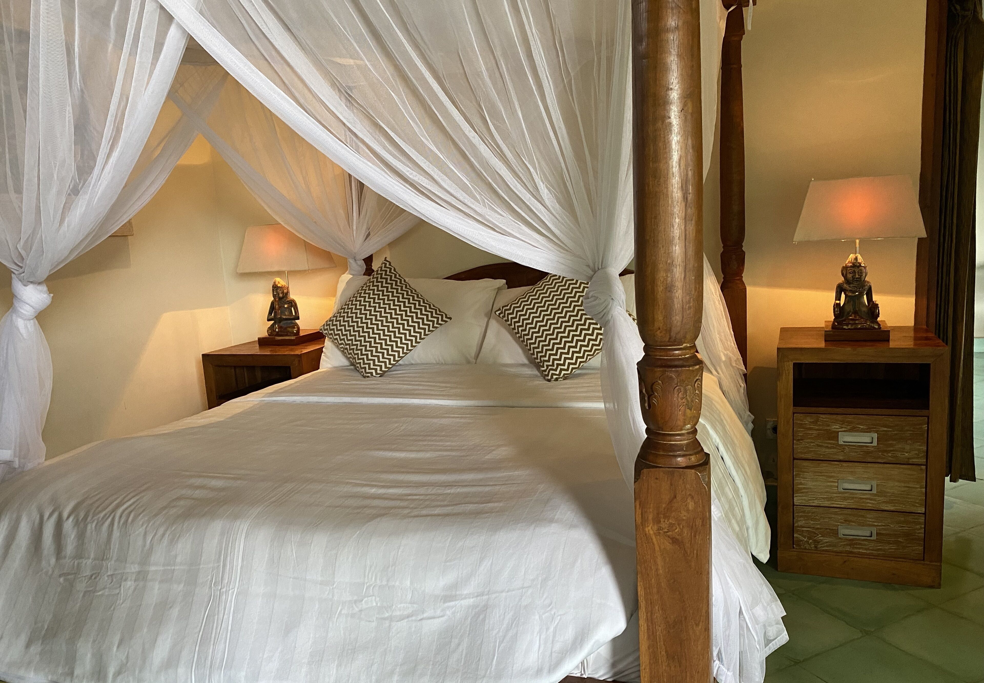 A four-poster canopy bed with white linens, two nightstands with lamps, and a wooden dresser with drawers.