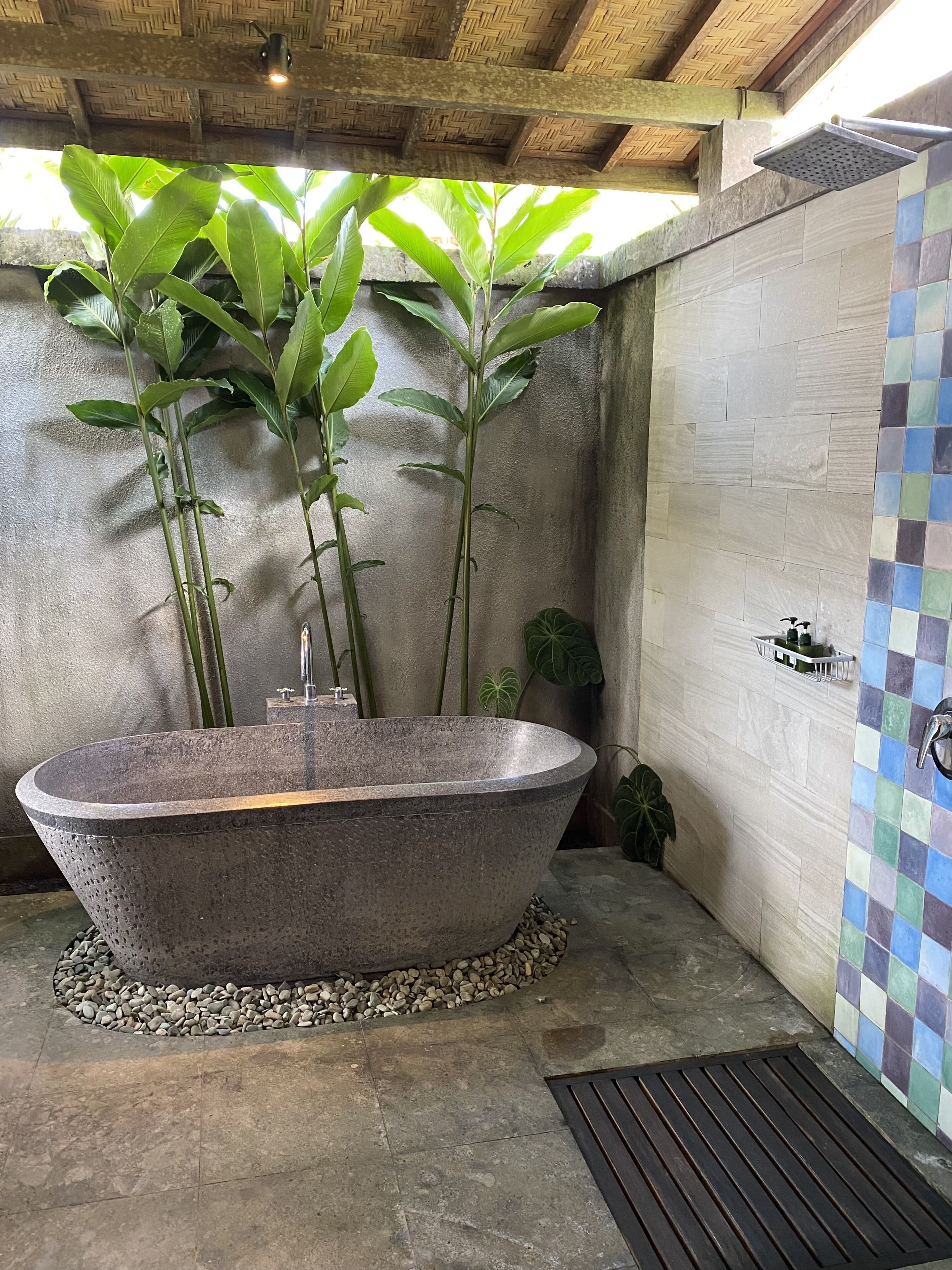 A stone bathtub in a bathroom with a tiled shower wall and a woven ceiling.