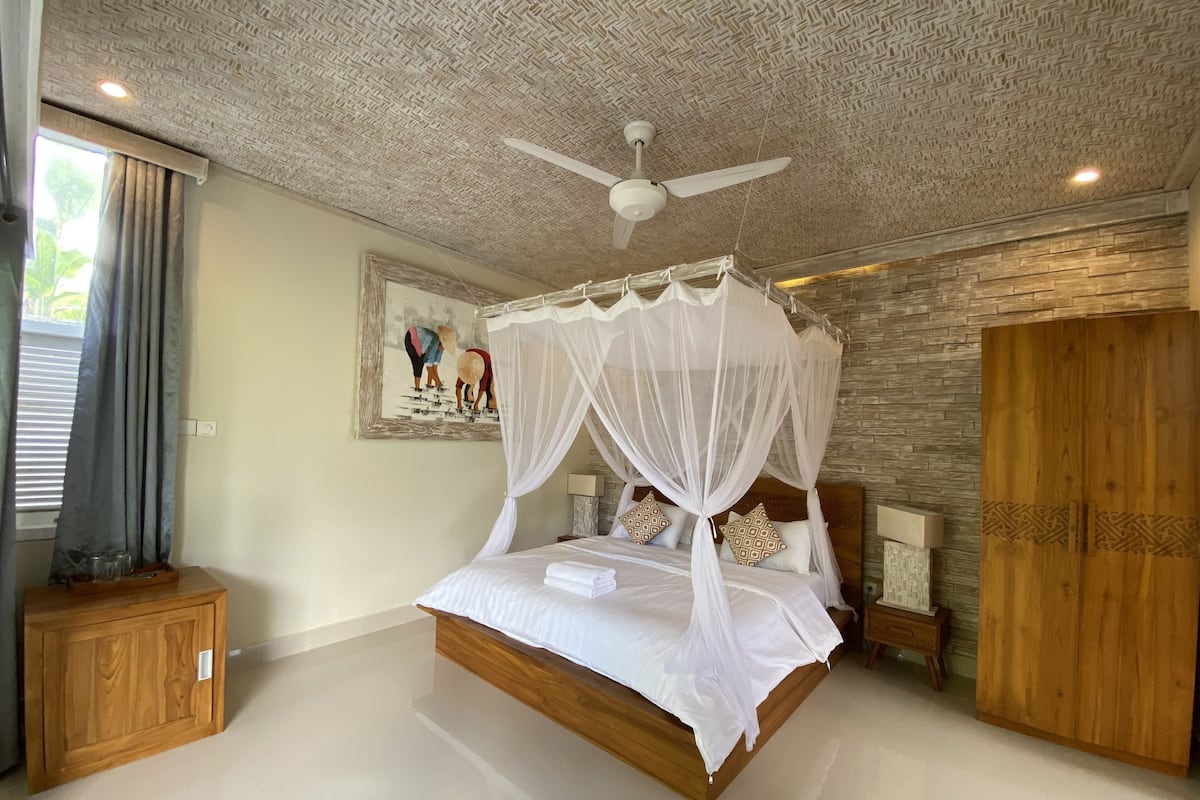 Un dormitorio con cama con dosel, ventilador de techo, armario de madera y un cuadro en la pared.
