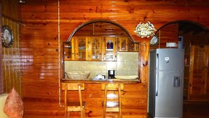 A wooden cabin interior with a kitchen area featuring wooden cabinets, a refrigerator, and a bar stool. - Little Bay Cabins (Little Bay)