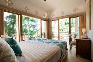 A spacious bedroom with a large bed, a wooden nightstand, and a view of a balcony with a chair and table. - Casa Mimba (Padangbai)