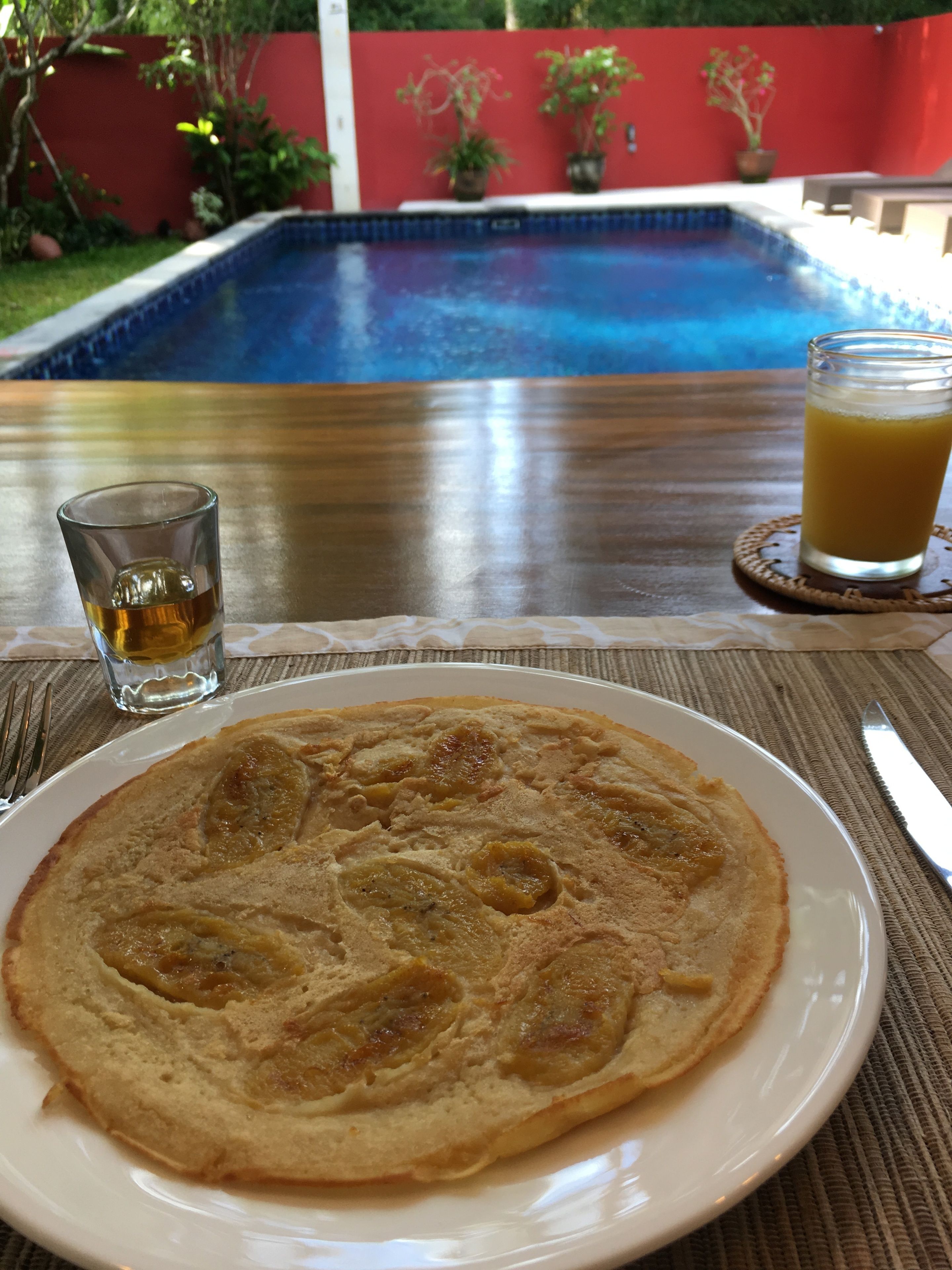 Un plato de desayuno con un panqueque, un vaso de jugo y un trago de licor sobre una mesa, con una piscina al fondo.