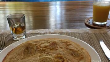 Un plato de desayuno con un panqueque, un vaso de jugo y un trago de licor sobre una mesa, con una piscina al fondo.